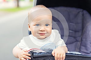 Cute caucasian blond curious baby boy sitting in stroller , smiling, looking and aspiring to something outdoor at city park.