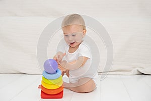 Cute caucasian blond smiling baby girl sitting alone on floor, playing with multi coloured pyramidion, light modern