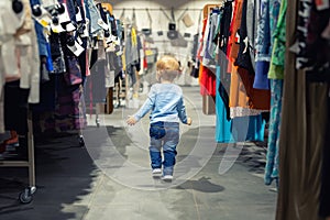 Cute caucasian blond toddler boy walking alone at clothes retail store between rack with hangers. Baby discovers adult shopping