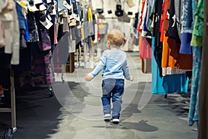 Cute caucasian blond toddler boy walking alone at clothes retail store between rack with hangers. Baby discovers adult shopping