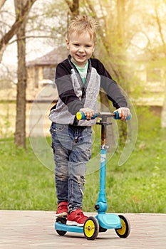 Cute caucasian boy with blue eyes and blond hair having fun at park with kick scooter.