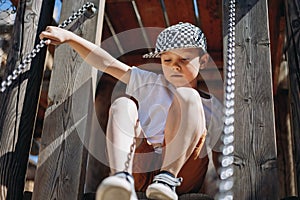 Cute caucasian boy in checkered  baseball cap and white tee shirt climbing down the ladder at the wooden playground.