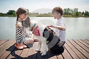 Cute caucasian boy and girl sitting by the river, smiling, cuddling the dog