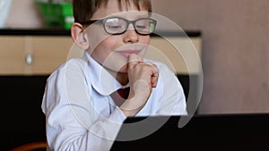 A cute caucasian boy in glasses 7 years old is studying at home doing homework sitting at a table in front of a gadget. Children a