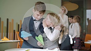 Cute Caucasian boy whispering on ear of friend with blurred classmates talking with teacher at the background