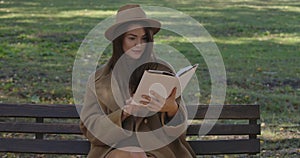 Cute Caucasian female student in elegant brown clothes sitting on the bench and turning book pages. Charming smart