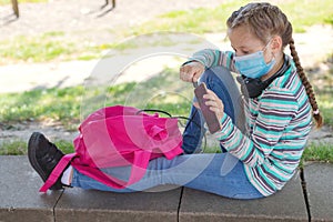 A cute Caucasian girl in a mask sits in the schoolyard with a backpack and a phone in her hands. Gadgets and kids. Rest from study