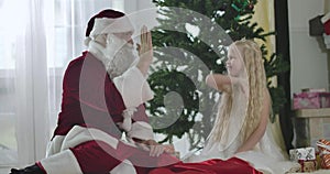 Cute Caucasian girl in white dress and crown sitting with Santa in front of New Year tree and giving him high five