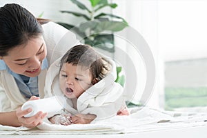 Cute Caucasian little toddler baby girl wear bathrobe after bathing is smiling and lying while mother apply  talcum powder on her