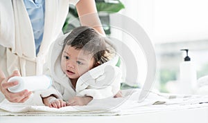 Cute Caucasian little toddler baby girl wear bathrobe after bathing is smiling and lying on towel. mom apply talcum powder on body