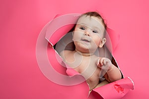 Cute Caucasian newborn baby boy peeks out of a hole in a paper pink background.