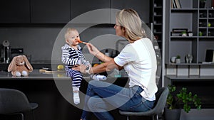 Cute Caucasian toddler sitting on tabletop as woman feeding baby with spoon. Portrait of pretty baby girl enjoying food