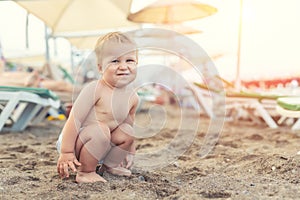 Cute caucasian toodler boy squatting alone on sandy beach between chaise-lounge. Adorable happy child having fun playing at
