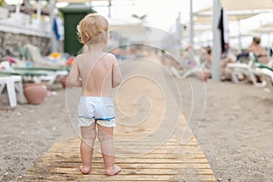 Cute caucasian toodler boy walking alone on sandy beach between chaise-lounge. Adorable happy child having fun playing