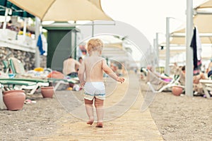 Cute caucasian toodler boy walking alone on sandy beach between chaise-lounge. Adorable happy child having fun playing at seaside