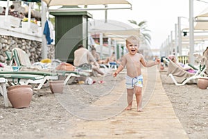 Cute caucasian toodler boy walking alone on sandy beach between chaise-lounge. Adorable happy child having fun playing at seaside