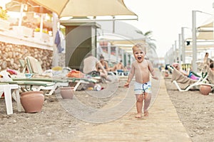 Cute caucasian toodler boy walking alone on sandy beach between chaise-lounge. Adorable happy child having fun playing at seaside