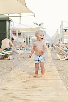 Cute caucasian toodler boy walking alone on sandy beach between chaise-lounge. Adorable happy child having fun playing at seaside