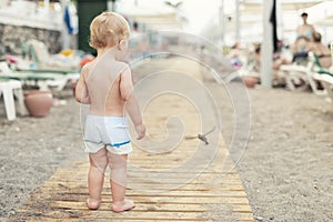 Cute caucasian toodler boy walking alone on sandy beach between chaise-lounge. Adorable happy child having fun playing at seaside