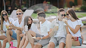 Cute caucasian woman feeding yummy pizza to her boyfriend on the background of their friends near poolside. Young people