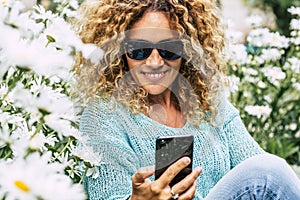 Cute caucasian woman using mobile phone outdoor sitting with daisies spring flowers around. Happy female people chat and send photo