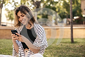 Cute caucasian young girl reads message on phone sitting outdoors during day.