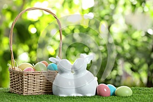 Cute ceramic Easter bunnies with wicker basket and dyed eggs on green grass against blurred background