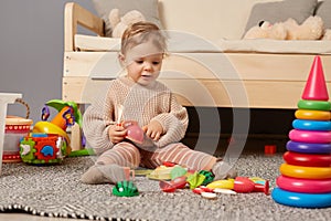 Cute charming baby girl wearing warm beige jumper sitting on floor near sofa and playing with toys alone at home, early
