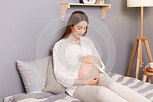Cute charming pregnant brown haired caucasian woman wearing white shirt sitting in bed touching holding her belly relaxing resting