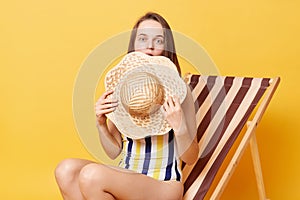 Cute charming surprised woman wearing striped swimming suit sitting on deck chair isolated over yellow background covering half of
