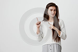 Cute cheerful brunette young female looking at camera and pointing forefingers away, showing something interesting and photo