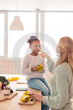 Cute cheerful girl holding pepper while helping mother cooking