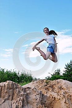 cute cheerful girl makes a jump in air against background of blue sky. Female athlete in white T-shirt and denim shorts performs