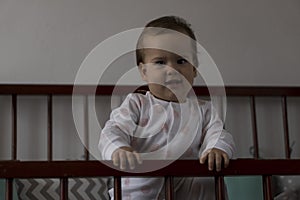 Cute cheerful little chubby baby girl sitting in grey baby crib after waking up from sleep in white and pink pajamas