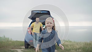 Cute cheerful little girl running along summer meadow on river bank with blurred father in car trunk at background