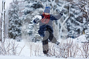 Cute, cheerful young boy in hat, blue jacket plays with snow, has fun, smiles, makes snowman in winter park.