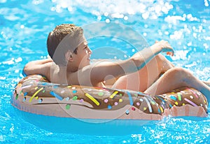 Cute child boy on funny inflatable donut float ring in swimming pool with oranges. Teenager learning to swim, have fun outdoor