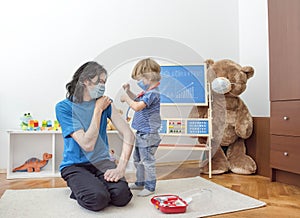 Cute child boy in home quarantine holding syringe with vaccine, playing doctor with dad wearing medical mask, role playing game