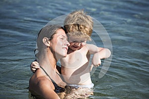 Cute child boy play with water in ocean, sea. Motherhood concept. Mother carries son in hands, hugs child. Mom and kid