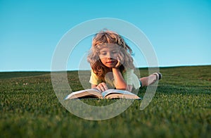 Cute child boy reading book outdoor on green grass field. Smart child reading book, laying on grass summer park on sky