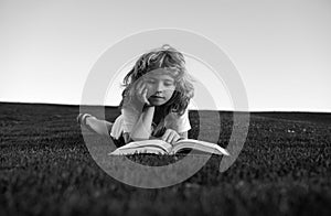 Cute child boy reading book outdoor on green grass field. Smart child reading book, laying on grass summer park on sky