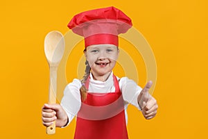 Cute child chef on a yellow background. A little girl holds a wooden spoon in her hands and shows her thumb up