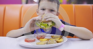 Cute child eating large hamburger at the table in fast food restaurant