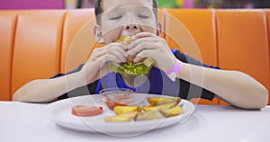 Cute child eating large hamburger at the table in fast food restaurant