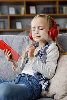 Cute child enjoy listening music lying on coach at home. Happy little girl wearing red headphones choosing favorite