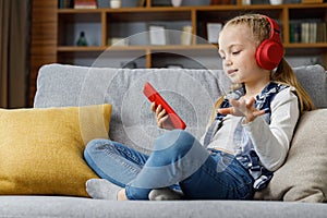 Cute child enjoy listening music lying on coach at home. Happy little girl wearing red headphones choosing favorite