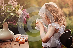 Cute child girl on cozy outdoor tea party in spring garden with bouquet of lilacs