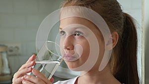 Cute child girl drinking clean transparent water from glass in kitchen at home