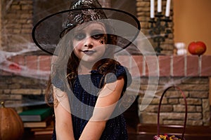 Cute child girl enchantress, in wizard& x27;s hat, sits against a cobweb-covered fireplace with spell books, smiles at