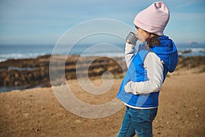 Cute child girl holds one hand in pocket, standing on the beach and drinking tea from a thermos cup, admiring the sea